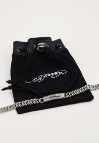 Pochette en toile noire avec une fermeture à cordon, dotée d'un bord supérieur en cuir et d'un bracelet en chaîne argentée avec un fermoir en métal. Logo "Ed Hardy" présent.