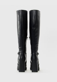Botas de cuero negras hasta la rodilla con tacones gruesos, cierres de cremallera en la parte posterior y correas en el tobillo con hebillas plateadas, mostradas desde la parte trasera.
