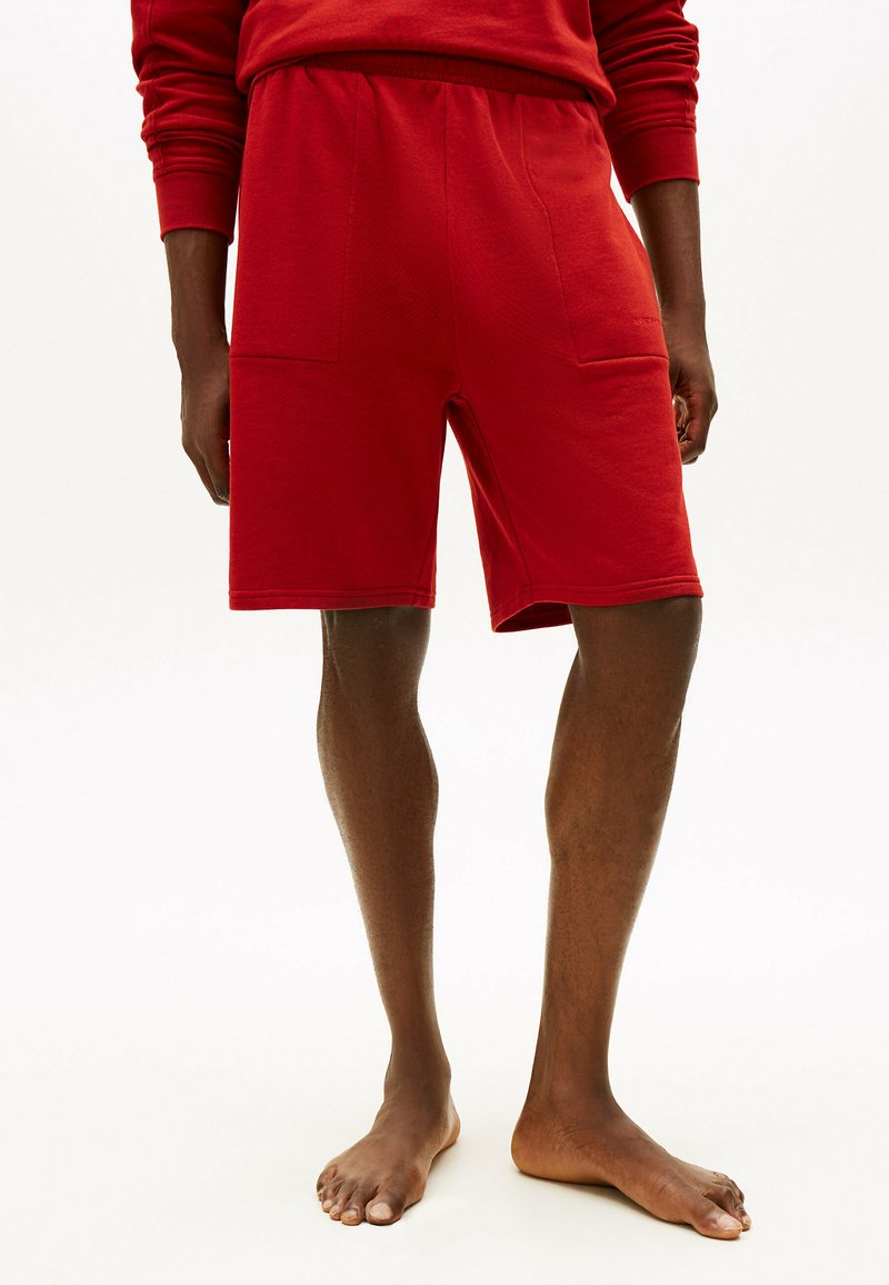 Personne portant un short rouge jusqu'aux genoux et un haut assorti à manches longues rouge, debout pieds nus sur un fond clair.