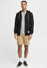 Sweat à capuche noir avec des cordons blancs, t-shirt gris, shorts beiges et baskets noires. La texture est lisse ; tenue décontractée et polyvalente.
