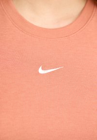 Rožinė banguota liemenė su baltu Nike swoosh logotipu, esantis ant krūtinės. Kaklo iškirpimas klasikinis, apvalus.