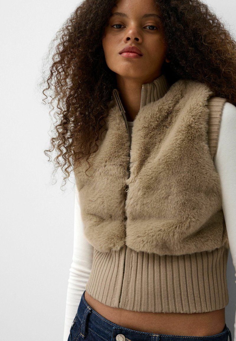 Beige gefälschtes Pelz-Weste mit geripptem Saum und hohem Kragen, ausgestattet mit einem Frontreißverschluss und ärmellosem Design, über einem weißen Longsleeve-Oberteil getragen.