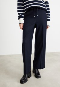 Marinblå vida byxor med dragsko i midjan och sidosfickor, kombinerade med svarta platåstövlar på en ljusgrå yta.