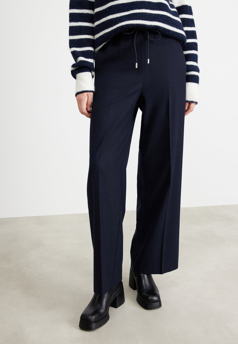 Marinblå vida byxor med dragsko i midjan och sidosfickor, kombinerade med svarta platåstövlar på en ljusgrå yta.