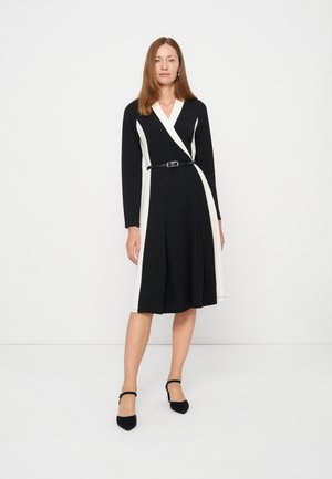 Femme portant une robe portefeuille noire et blanche à manches longues avec ceinture, debout sur un fond blanc uni, portant des talons noirs à bride à la cheville.