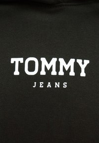 Čierna mikina s bielym logom s tučnými písmenami "TOMMY" nad "JEANS." Hladká textúra s priliehavým strihom.