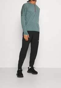 Long-sleeve teal sports shirt with a red accent and textured design, paired with black athletic pants and black sneakers.