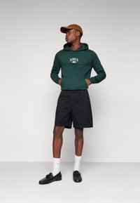 Sudadera verde con bolsillo delantero y texto blanco "DENVER", pantalones cortos negros, calcetines blancos y mocasines negros. Una gorra marrón completa el conjunto.