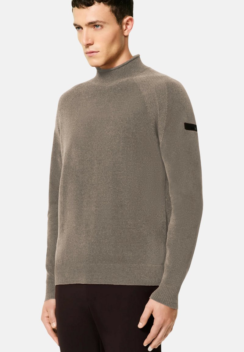 Uomo con un maglione a collo alto color taupe, maniche lunghe e pantaloni neri, in piedi davanti a uno sfondo grigio chiaro uniforme.