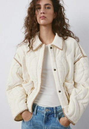 Donna con capelli ricci che indossa una giacca trapuntata color crema con rifiniture marroni, maglietta bianca e jeans blu, mani nelle tasche della giacca, sfondo neutro.