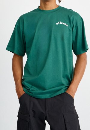 Person trägt ein grünes ellesse T-Shirt mit kurzen Ärmeln und schwarze Cargo-Hosen, Hände in den Taschen, vor einem schlichten Hintergrund.