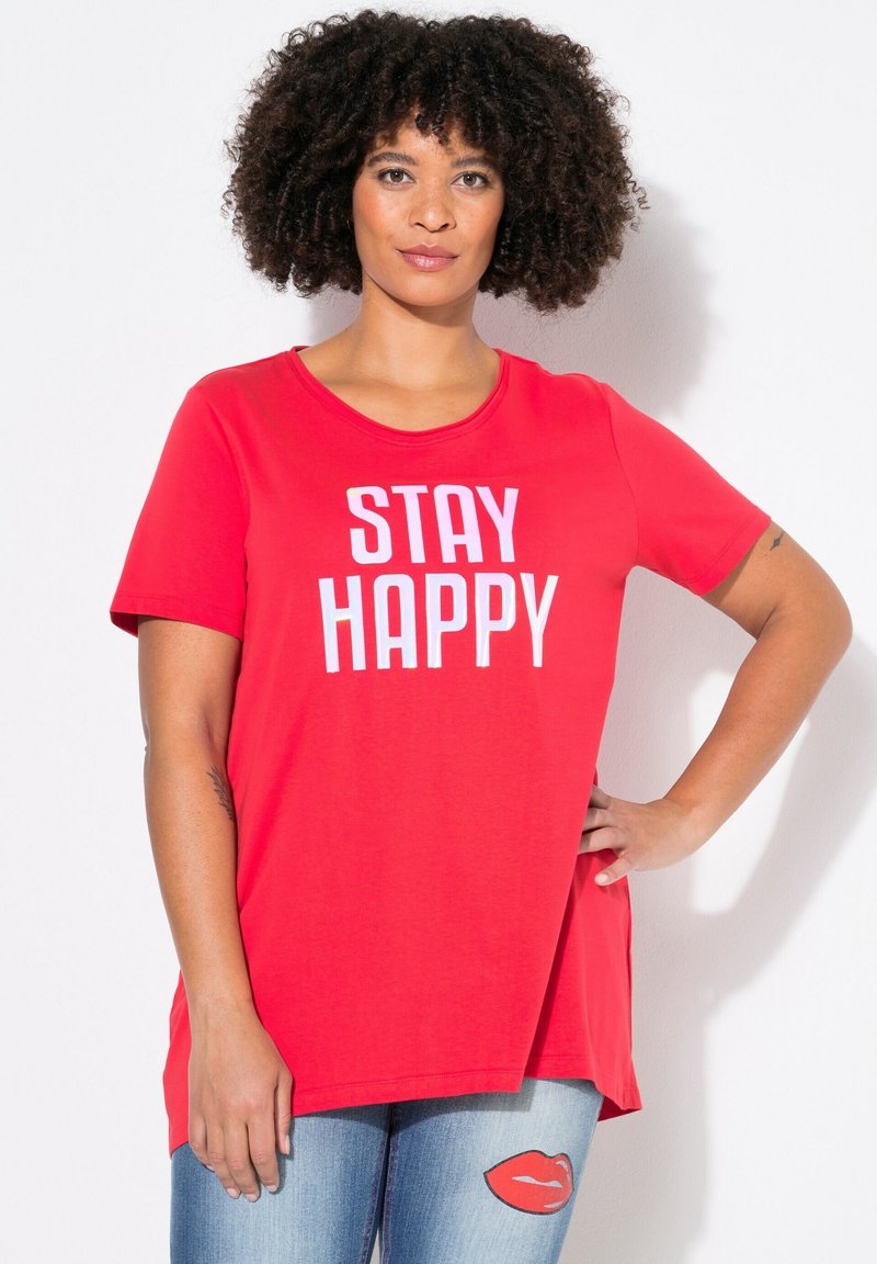 Rode T-shirt met korte mouwen en de tekst "STAY HAPPY" in het wit. Zachte stof, ronde halslijn, licht langere achterkant, gecombineerd met lichtblauwe jeans met lippenontwerp.