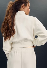 Felpa bianca corta con maniche lunghe e collo alto. Il materiale sembra morbido e strutturato, abbinato a pantaloni con elastico in vita dello stesso colore.