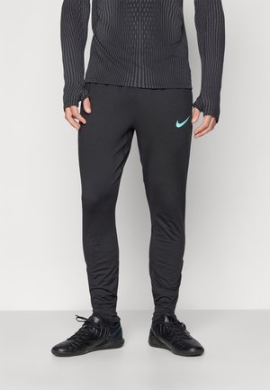 Man draagt zwarte Nike sportbroek met blauwgroen logo en zwarte sneakers tegen een witte achtergrond.