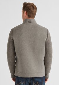 Grauer Wollpullover mit hohem Kragen, strukturierten Rippärmeln und glattem Körper. Besitzt ein dezentes Logodetail am Kragen.