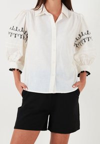 Chemisier blanc à boutons avec broderie noire sur des manches bouffantes, doté d'un tissu texturé et de shorts noirs contrastants.