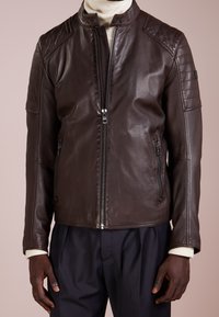 Brun läderjacka med dragkedja fram, kviltade axel detaljer och sidofickor. Har en stå krage och en figurnära design.