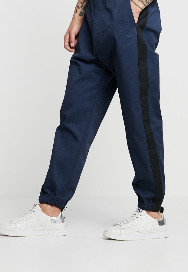 Personne portant un pantalon de jogging bleu marine avec des bandes latérales noires et des baskets blanches, debout avec une main dans la poche.