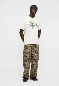 Νέος άνδρας που φοράει κρεμ μπλουζάκι με πράσινο κείμενο, καμουφλαρισμένο παντελόνι cargo, μαύρα παπούτσια και ελιά πράσινο πλεκτό σκουφί, στέκεται με τα χέρια πίσω από την πλάτη.