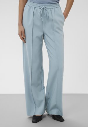 Pantalon large bleu clair avec taille élastique et cordon, porté avec des chaussures noires pointues et un haut rentré.