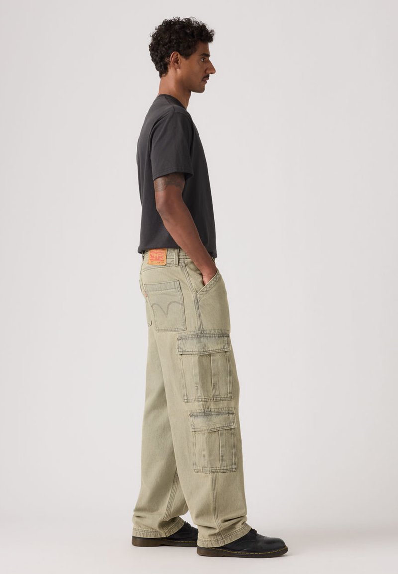 Hellgrüne Cargo-Hose mit mehreren Taschen und Ziernähten, kombiniert mit einem schwarzen Kurzarmhemd und schwarzen Schuhen.