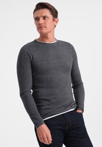 Graues Langarmshirt mit strukturiertem Strickmuster, ausgestattet mit einem weißen Rundhalsausschnitt und einer taillierten Passform, kombiniert mit dunklen Jeans.