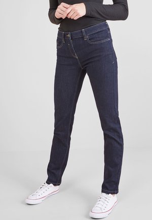 Person trägt dunkelblaue Slim-Fit-Jeans, schwarzen Langarm-Top und weiße Sneaker mit niedrigem Schaft, steht mit verschränkten Händen.