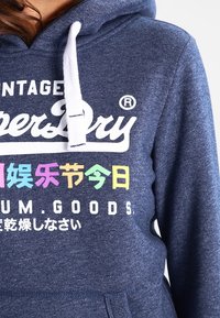 Laivastonsininen huppari, jossa on kiristysnaru, etutasku ja monivärinen teksti. Materiaalissa on teksturoitu pinta. Brändin logo näkyvällä paikalla.