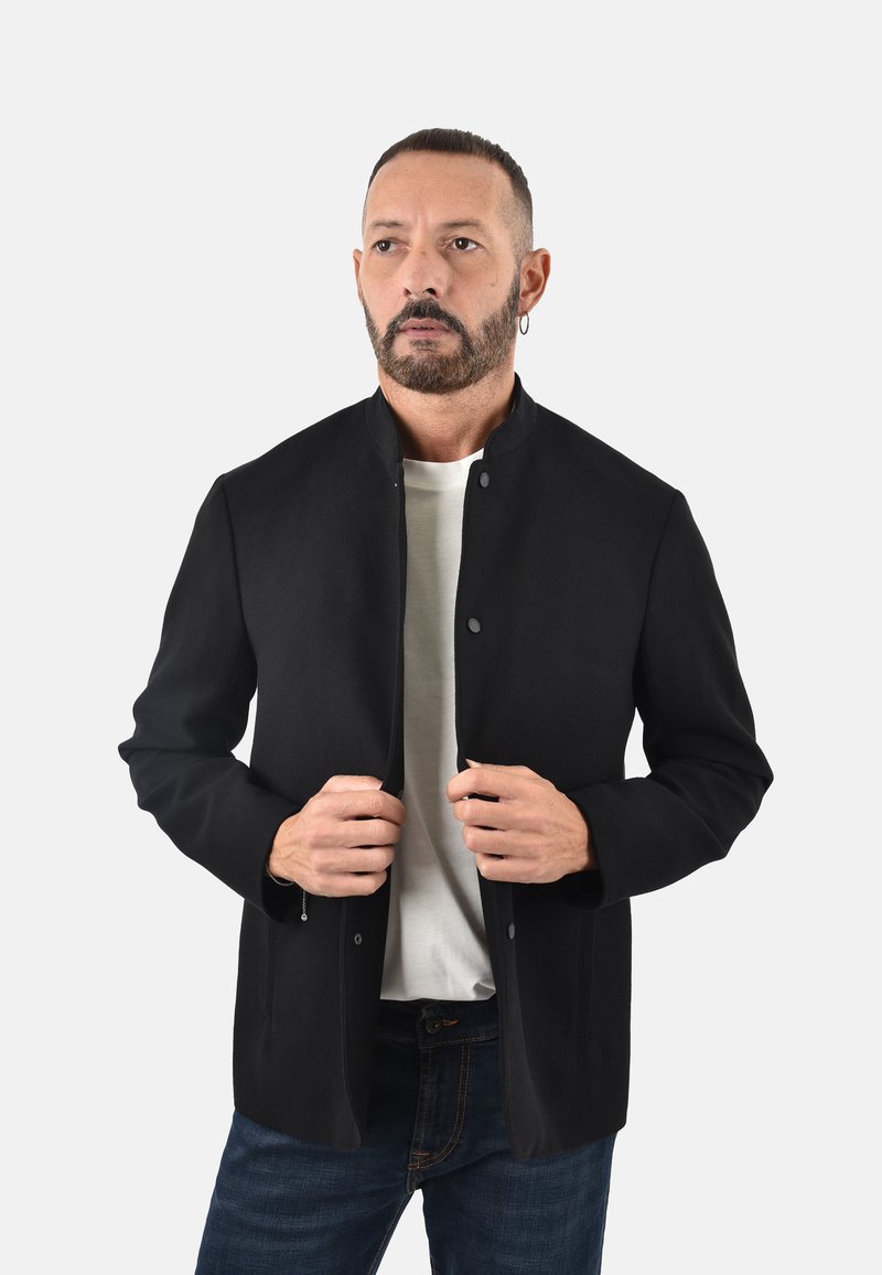 Giacca nera su misura con colletto rialzato, bottoni a pressione e maniche lunghe. Indossata sopra una camicia bianca con una texture di tessuto liscia.