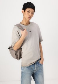 T-shirt oversize gris clair avec un petit logo noir sur la poitrine, porté avec un jean en denim bleu et un sac à épaule gris doux.