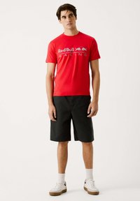 Rød T-shirt med "Red Bull Racing" tekst i grå, parret med sorte shorts. Modellen bærer hvide sokker og sneakers, og står foran en ensfarvet baggrund.