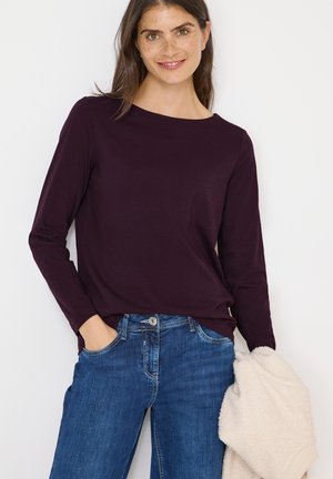 Långärmad, rundhalsad topp i djuplila, matchad med blå denimjeans. En krämfärgad, fluffig jacka hålls i armen.