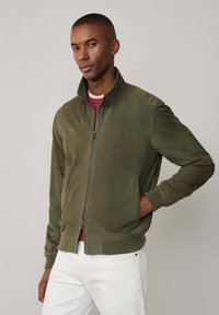 Veste bomber en daim vert olive avec un col haut, fermeture éclair, poches latérales et poignets côtelés, portée sur une chemise rouge et un pantalon blanc.