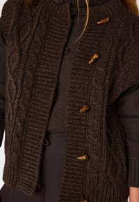 Cardigan a maglia grossa in lana marrone scuro, con dettagli a costine, bottoni a chiusura e una texture spessa. Indossato sopra un dolcevita marrone.