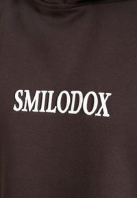 Sweatshirt marron avec le texte brodé en blanc "SMILODOX", doté d'un tissu lisse et texturé et d'un design simple et décontracté.