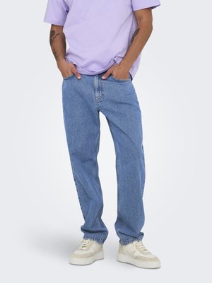 Person wearing light purple t-shirt, loose blue jeans with hands in pockets, and beige sneakers, standing against a plain background.