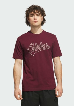 Young man with curly hair wearing a maroon Adidas t-shirt with white script logo, standing against a plain light background.