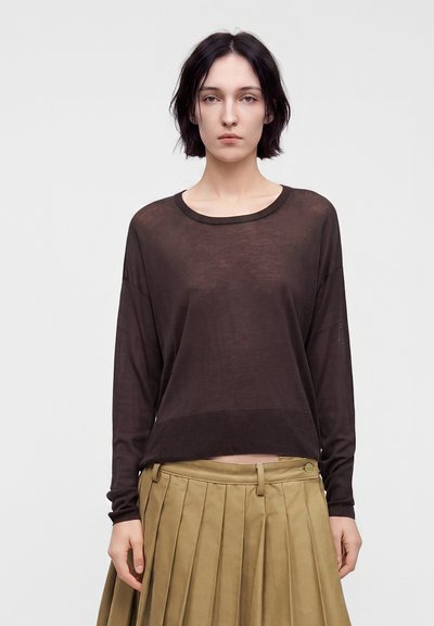 Femme aux cheveux courts et foncés, portant un haut transparent à manches longues marron foncé et une jupe plissée beige taille haute sur un fond uni.