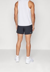 Hombre vestido con una camiseta sin mangas blanca, pantalones cortos atléticos negros, calcetines blancos y zapatillas blancas, de espaldas a la cámara, sobre un fondo liso.