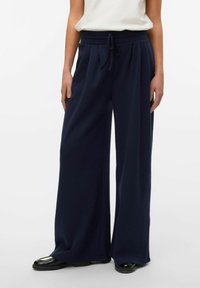 Pantaloni a gamba larga blu navy con vita regolabile, pieghe e tessuto liscio. Abbinati a una maglietta bianca e scarpe nere lucide.