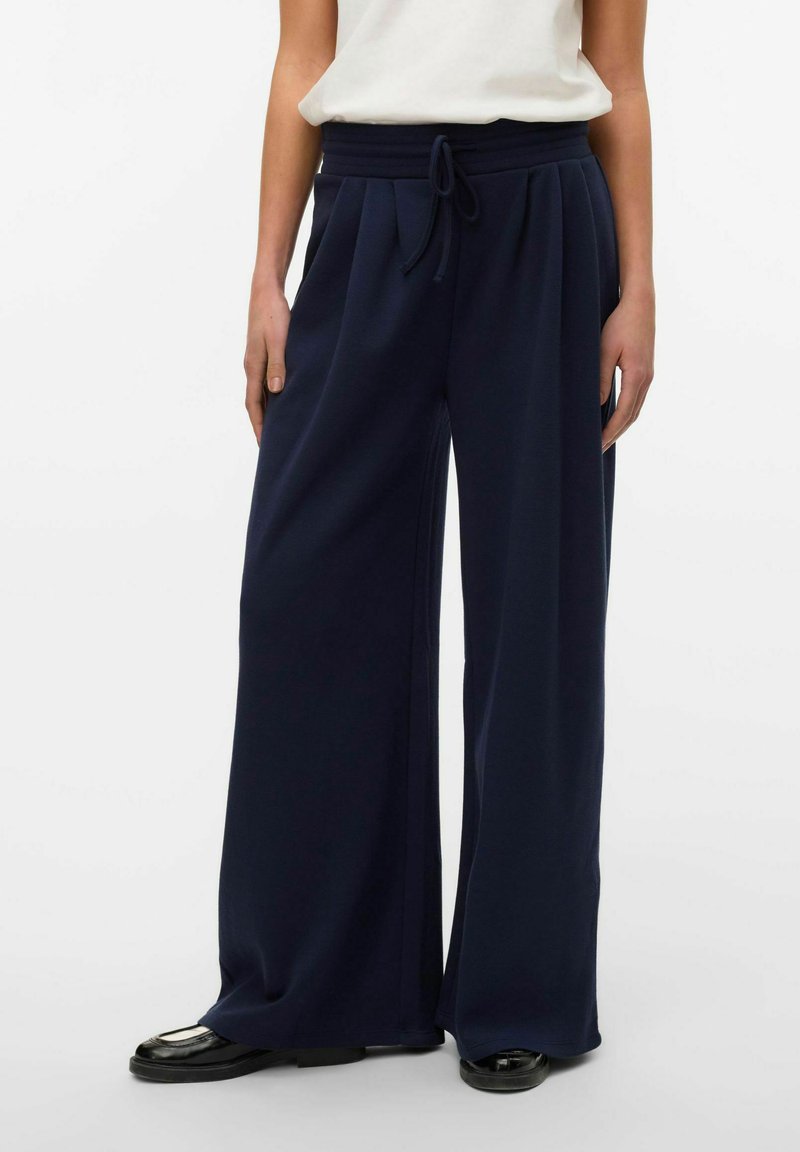 Pantaloni a gamba larga blu navy con vita regolabile, pieghe e tessuto liscio. Abbinati a una maglietta bianca e scarpe nere lucide.