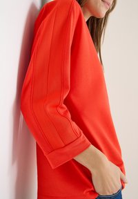 Punainen pitkähihaispaida, jossa on ribbikuosi ja käännetyt hihat. Kangas vaikuttaa pehmeältä ja jämäkältä, korostaen rentoa siluettia.