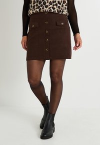 Minifalda de pana marrón con botones en el frente, dos bolsillos con solapa y adornos de hardware dorado. Llevada con botas negras hasta el tobillo y medias.