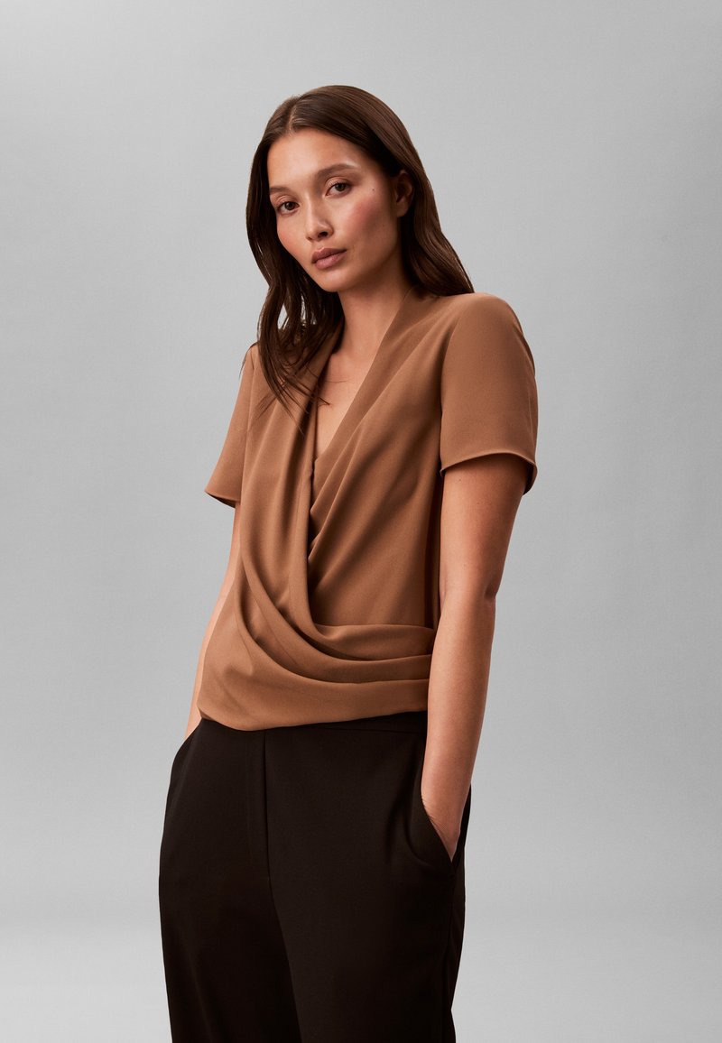 Νεαρή γυναίκα με καστανά μαλλιά φοράει καφέ κοντομάνικη μπλούζα με draped λεπτομέρειες και μαύρο παντελόνι, στέκεται με τα χέρια στις τσέπες μπροστά σε γκρι φόντο.