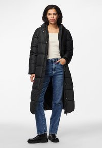 Casaco puffer longo preto com capuz, textura acolchoada, botões de pressão e rachas laterais, usado sobre uma blusa creme e jeans de denim azuis. Botas ankle pretas.