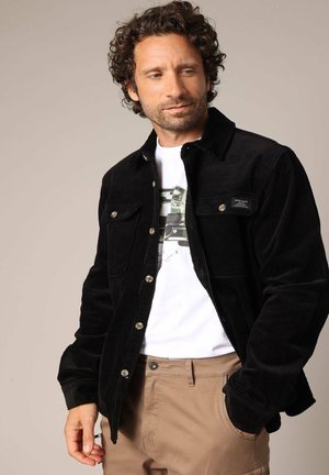 Veste en velours côtelé noir avec un design à col, dotée de deux poches poitrine et de boutons en argent, portée sur un t-shirt graphique blanc.