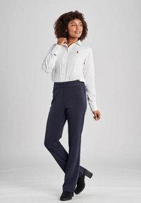 Witte blouse met een logo, gecombineerd met marineblauwe broek met elastische taille. Model draagt zwarte hakken. Eenvoudig ontwerp, gladde textuur.