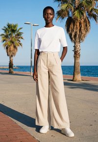 Femme debout sur la promenade en bord de mer portant un t-shirt blanc, un pantalon large beige et des baskets blanches ; palmiers et océan en arrière-plan.