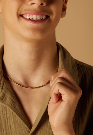Gouden ketting die op de nek rust, gedragen met een olijfgroen kledingstuk met kraag. De hand houdt de kraag vast. Soepele textuur zichtbaar.