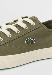 Baskets en toile vert olive avec des lacets blancs, arborant un petit logo de crocodile brodé et une semelle en caoutchouc crème.